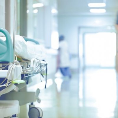 A hospital corridor with a trolley bed on one side, and an out of focus outline of a hospital worker in green scrubs on the other. 